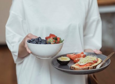 Person holding a bowl of fresh berries and a plate with avocado toast, displaying a healthy breakfast spread.